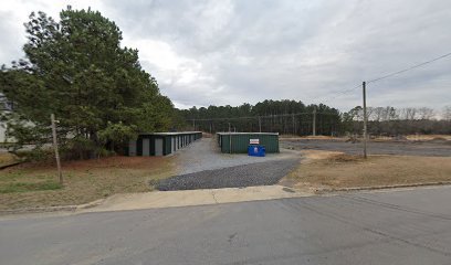 Entrance view down a gravel lane between storage buildings with pine trees behind